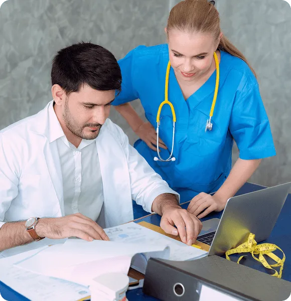 Man in white coat and woman in blue scrubs reviewing documents beside a laptop and folder