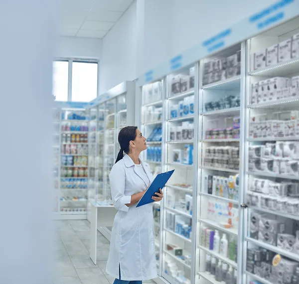 pharmacy image 59477 webp Pharmacist in a white coat holding a blue clipboard and looking up at shelves of boxed medicines