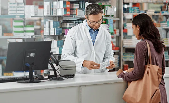 pharmacy image 59479 webp Pharmacist in a white coat and glasses holds a medicine box at a counter while a customer with a tote bag waits