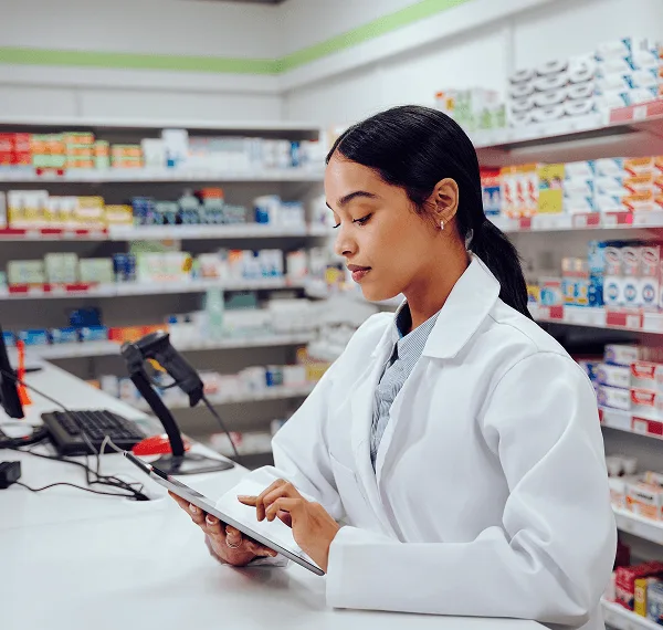 pharmacy image 59481 webp Pharmacist in a white coat uses a tablet at a counter with medicine shelves and a barcode scanner behind