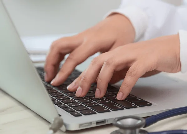 physiatry image 59466 webp Two hands typing on a laptop keyboard with a stethoscope resting beside it