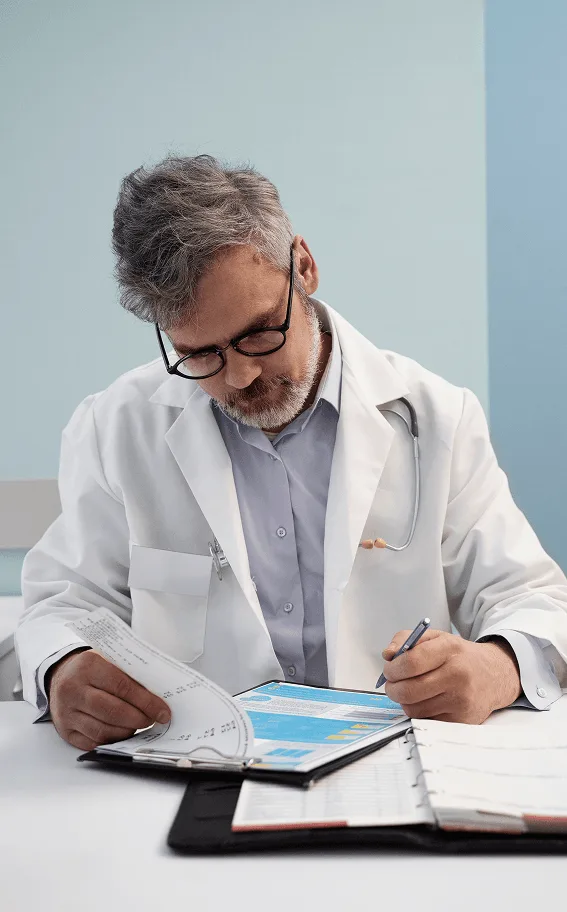 Doctor in a white coat with glasses reviewing papers and writing notes on a clipboard