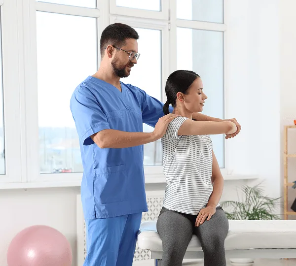 Physical therapist in blue scrubs guiding a seated patient through a shoulder stretch