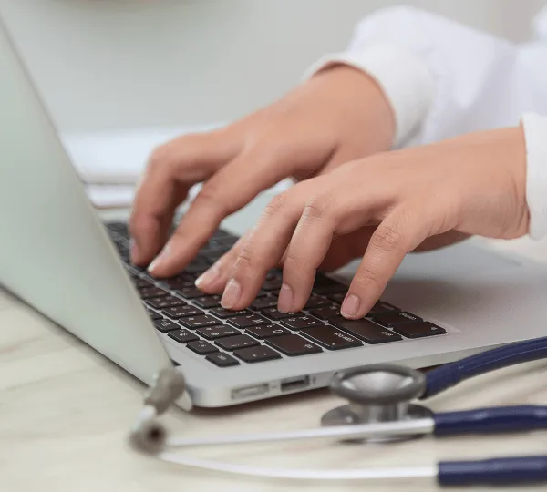 Hands typing on a laptop keyboard with a stethoscope lying on the desk beside it