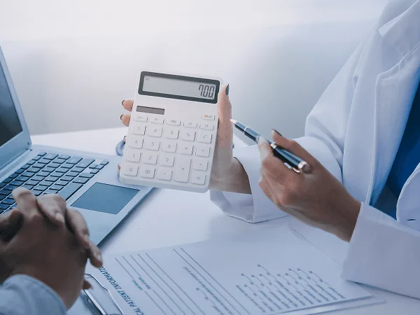 physician image 59443 webp Hand holding a white calculator beside a pen, with a laptop and patient information form on a desk