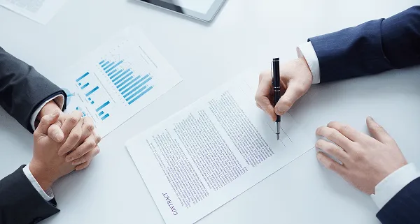 physician image 59445 webp Overhead view of a person signing a contract beside a printed bar chart while another waits