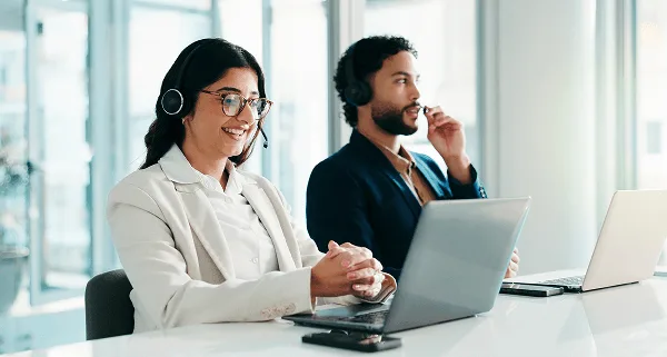 physician image 59446 webp Man and woman wearing headsets at a bright office desk with laptops, woman smiling