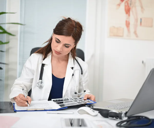 plastic-surgery image 59431 webp Female doctor in a white coat writing notes on a clipboard with an X-ray film at her desk