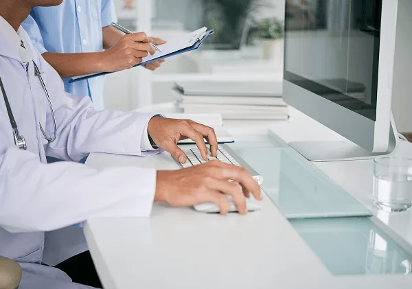 plastic-surgery image 59434 webp Clinician in a white coat using a keyboard and mouse while a colleague writes on a clipboard