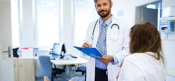 Doctor with a stethoscope holds a blue clipboard and talks with a seated patient in a clinic