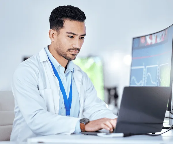 Man in a white coat with a lanyard works on a laptop beside a monitor showing charts