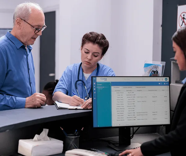 Nurse writes in a notebook at a reception desk while a patient stands beside a computer monitor