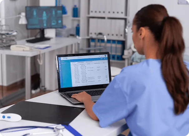 Nurse in blue scrubs types on a laptop showing a patient list in a medical office