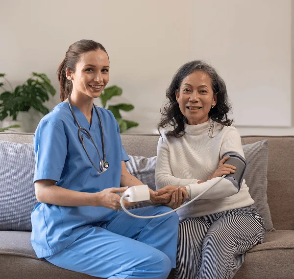 primary-care image 59412 webp Smiling nurse in blue scrubs with stethoscope taking an older woman's blood pressure on a sofa