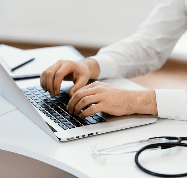 primary-care image 59413 webp Close view of hands typing on a laptop with a stethoscope lying on a white desk