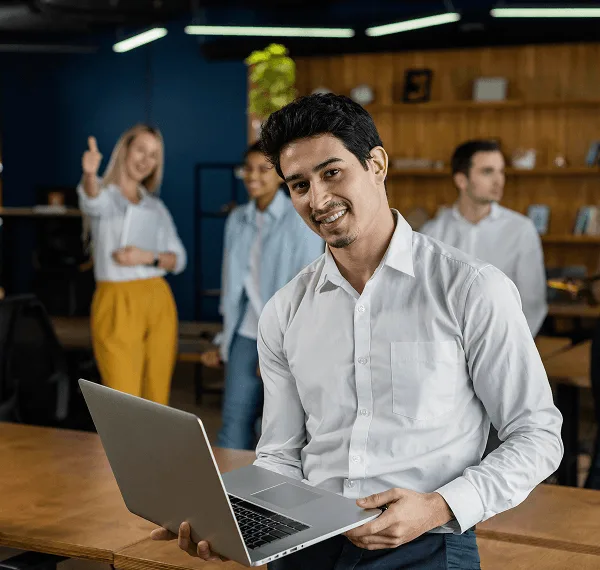 primary-care image 59414 webp Smiling man in white shirt holding a laptop in an office while colleagues stand blurred behind him