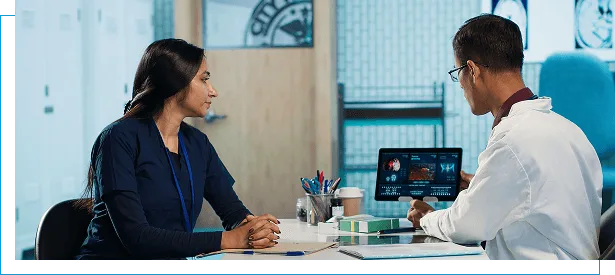 primary-care image 59415 webp Woman in dark top and a man in white coat sit talking over a tablet showing scans