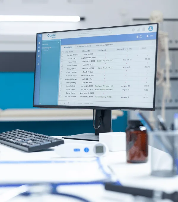 Desktop monitor showing a patient list on a clinic desk with a keyboard, thermometer and medicine bottle