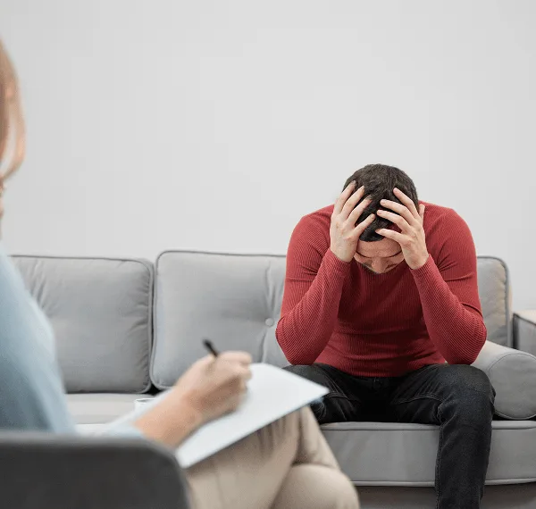 Man in a red sweater sits on a sofa with his head in his hands while someone takes notes