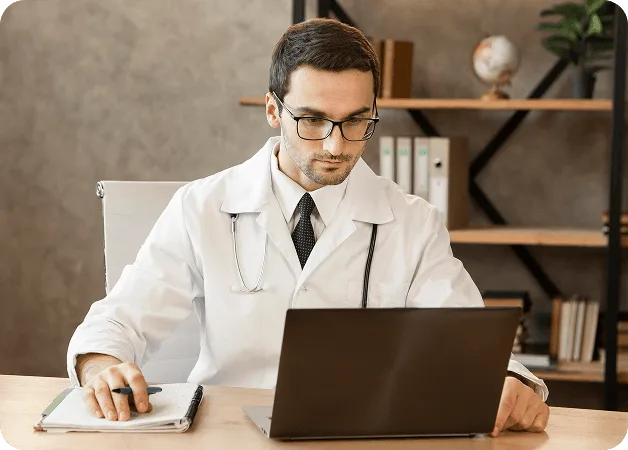 Man in a white coat and glasses writes in a notebook beside an open laptop at a wooden desk