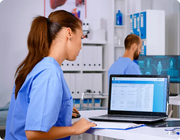 Woman with a ponytail in blue scrubs works at a laptop showing a data table while a bearded man stands behind her