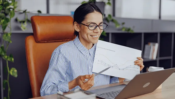 Smiling woman with glasses holding up a line chart printout in front of a laptop