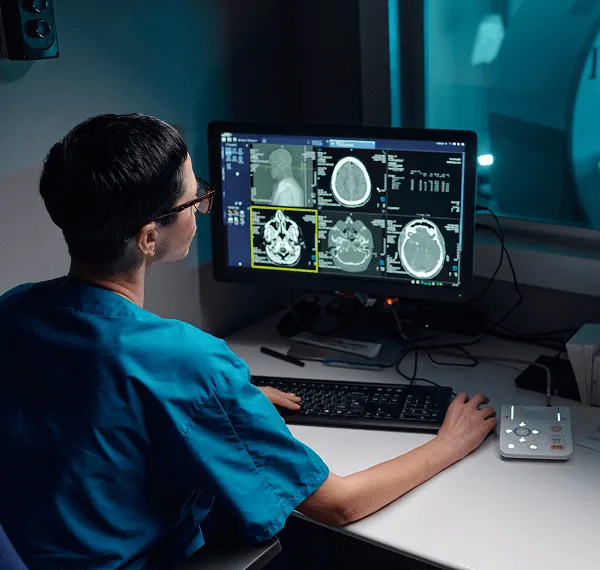 Radiology staff member in teal scrubs reviews brain scan images on a computer monitor