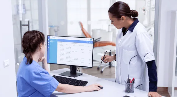 Two medical staff members look at a computer screen displaying a table of records in a clinic