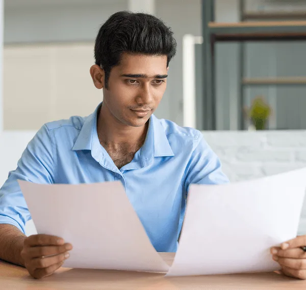 Man in a blue shirt looks at two sheets of paper while seated at a desk