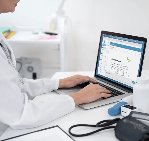 Clinician types on a laptop showing a medical history form beside a blood pressure cuff and health devices