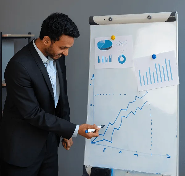 Man in a dark suit draws a rising line graph on a whiteboard with charts pinned to it