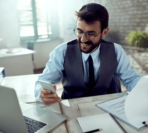 Bearded man with round glasses smiling at his phone at a desk with laptop and papers