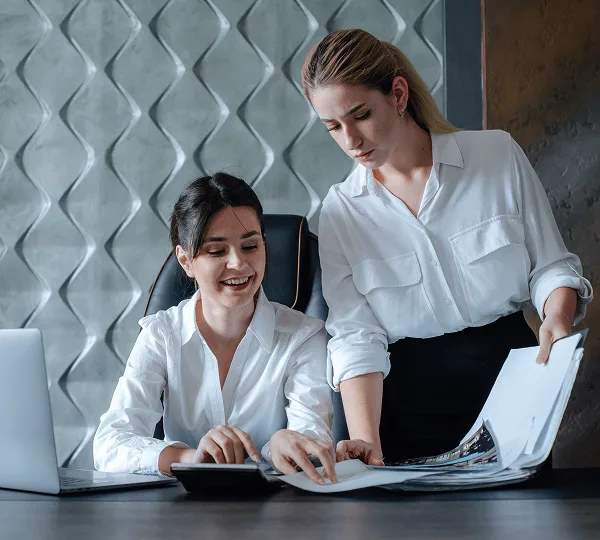Two women in white blouses reviewing documents and a calculator at a desk with a laptop