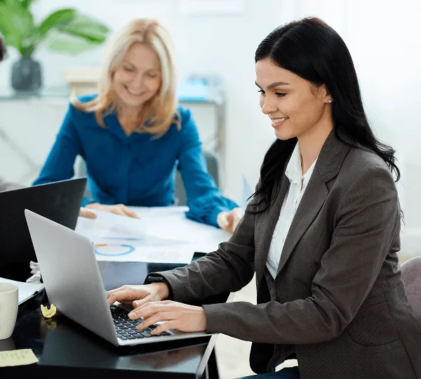 Two smiling office professionals working at a table, one typing on a laptop