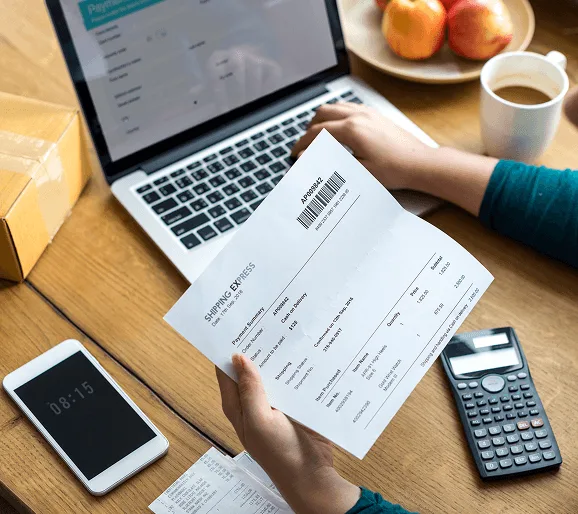 Hands holding a printed shipping invoice, with a laptop, calculator, phone and coffee cup on a wooden table