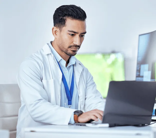 Doctor in a white coat with a lanyard typing on a laptop at a desk