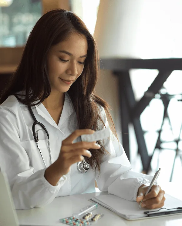 Woman in a white coat with stethoscope holds a pill blister pack while writing notes