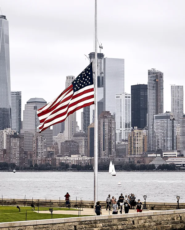American flag at half-staff on a pole with a city skyline across the water behind