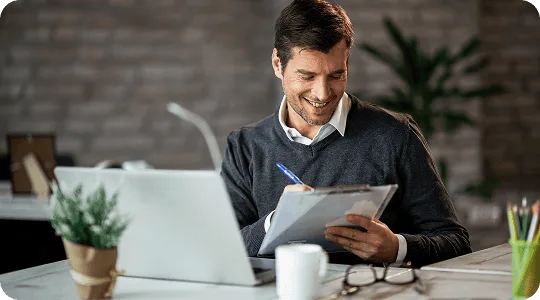 Smiling man writing on a clipboard at a desk with a laptop and coffee mug