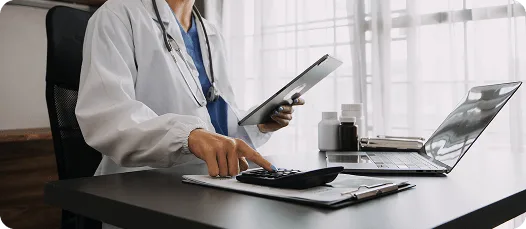 Doctor at a desk using a calculator and holding a tablet, with a laptop nearby