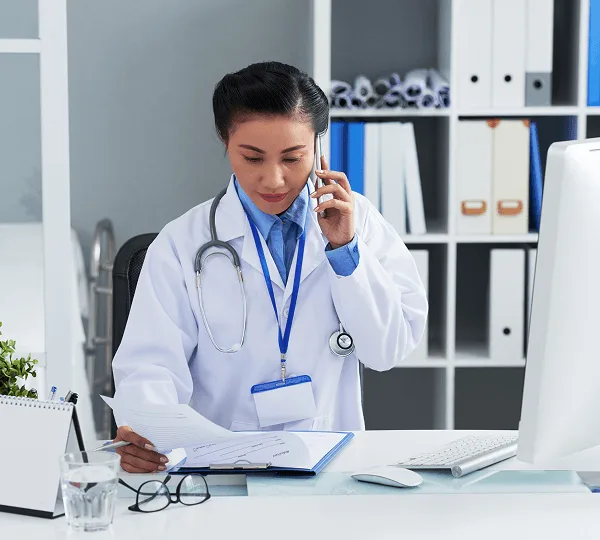 telehealth image 59253 webp Doctor in a white coat on a phone call, reviewing papers at a desk with a computer