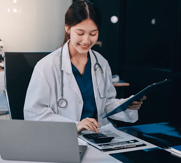 telehealth image 59254 webp Smiling woman in a white coat holding a clipboard while using a calculator at a desk