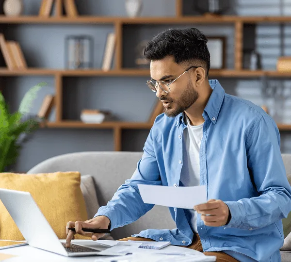 teleradiology image 59234 webp Man with glasses in a blue shirt holding a paper and typing on a laptop at home