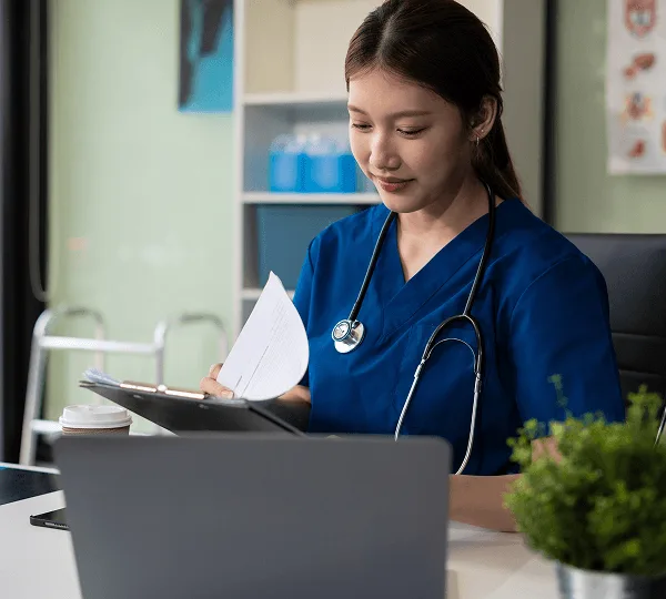 Nurse in blue scrubs with a stethoscope reviewing papers on a clipboard in front of a laptop