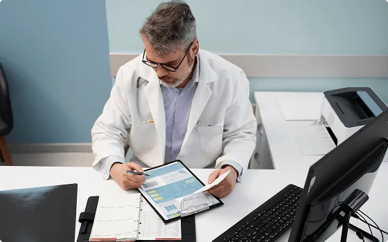 Man with glasses and gray hair in a white coat reviewing a chart at a desk with monitor and printer
